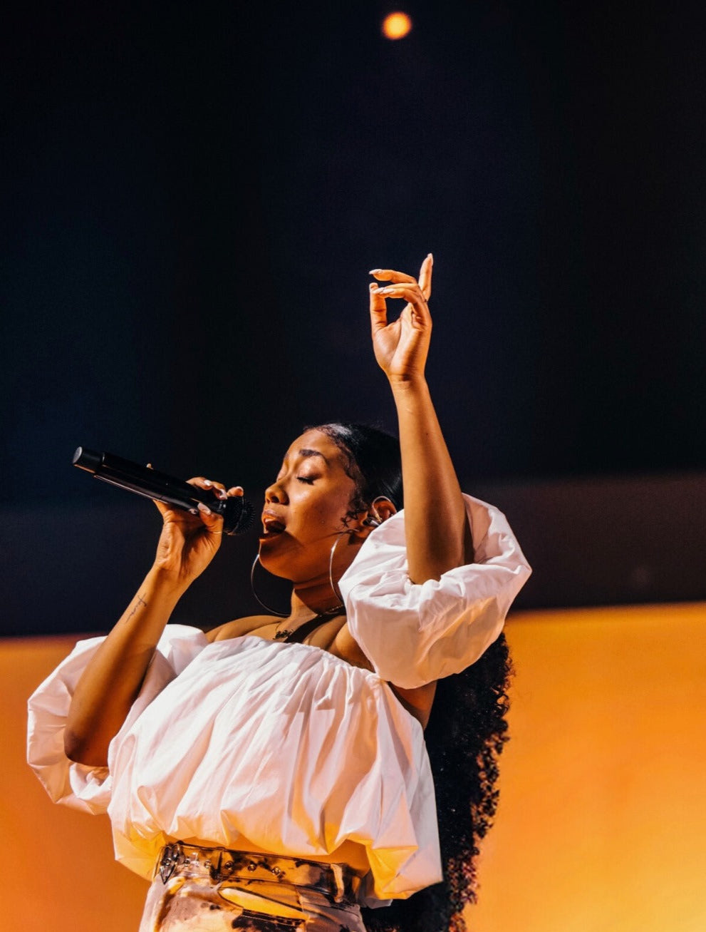 Ilaisa performing live indie-pop, singing into a mic with one hand raised, wearing a white off-shoulder top on stage